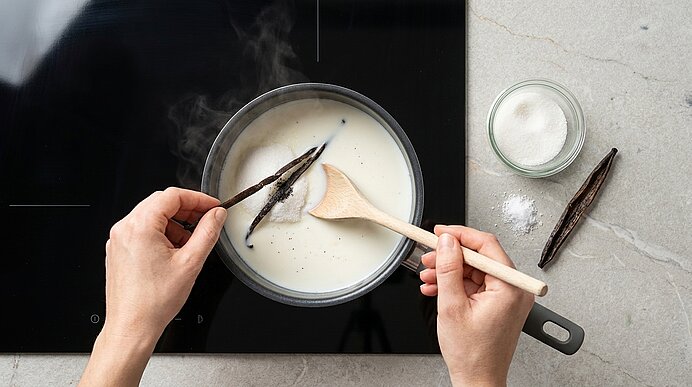 Hände halten Vanilleschoten über einem Topf mit heißer Milch auf dem Kochfeld; ein Holzlöffel rührt, Zucker im Glas daneben.