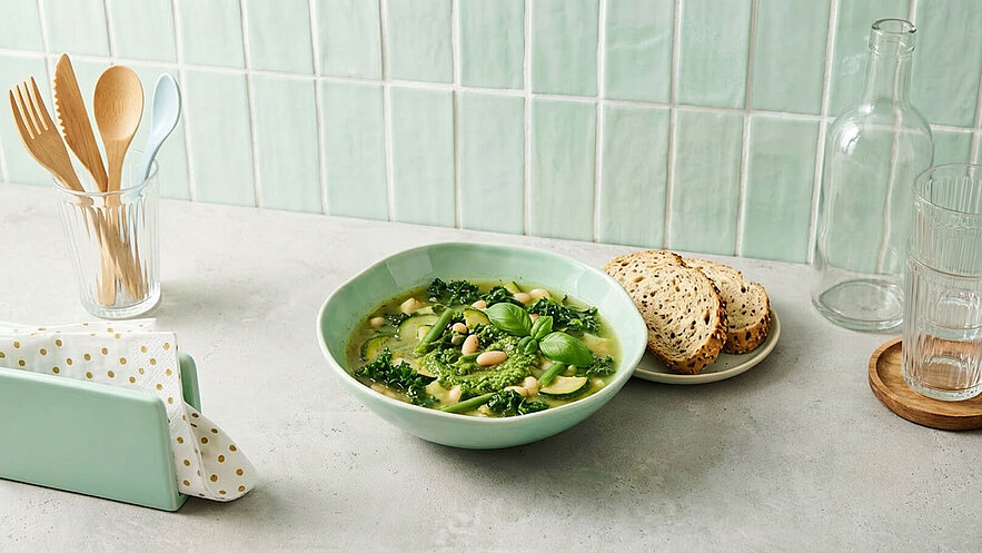Eine grüne Schüssel mit einer Gemüsesuppe, die Zucchini, Grünkohl und weiße Bohnen enthält, ist auf einem Tisch platziert. Daneben liegt ein Teller mit mehreren Scheiben Vollkornbrot. Im Hintergrund sind ein Glas mit Besteck und andere Esswaren sichtbar.