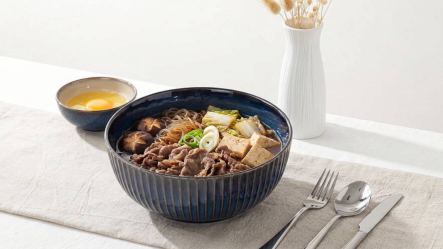 Eine Schüssel mit Ramen-Nudeln, belegt mit zartem Fleisch, Tofu, Frühlingszwiebeln und Gemüse. Daneben steht eine Schale mit einem rohen Ei und eine Vase mit Trockenblumen. Der Tisch ist einfach gedeckt mit Besteck und einer Tischdecke.
