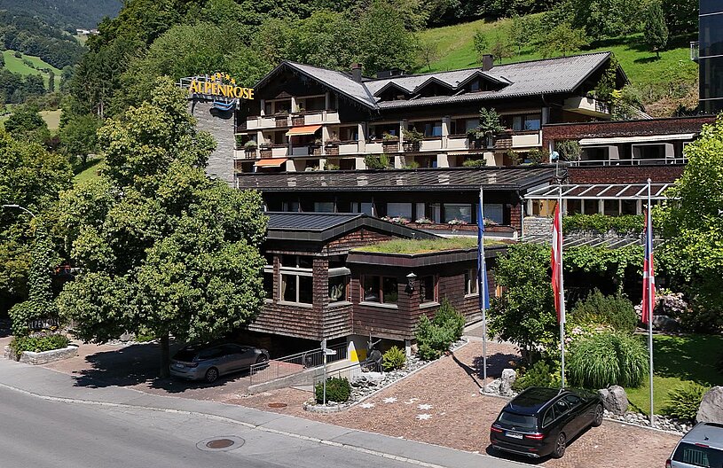 Ein modernes Hotelgebäude mit Holzverkleidung und mehreren Stockwerken, umgeben von grünen Bäumen und Wiesen. Vor dem Hotel stehen Autos geparkt. Im Hintergrund sind sanfte Hügel zu sehen. Ein Schild mit der Aufschrift „Alpenrose“ ist sichtbar.