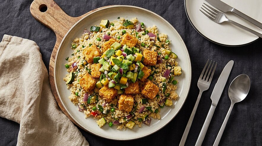 Teller mit Couscous-Salat, gewürfeltem Gemüse und Kräutern, belegt mit knusprig gebratenen Tofuwürfeln und frischer Gurken-Mango-Salsa.