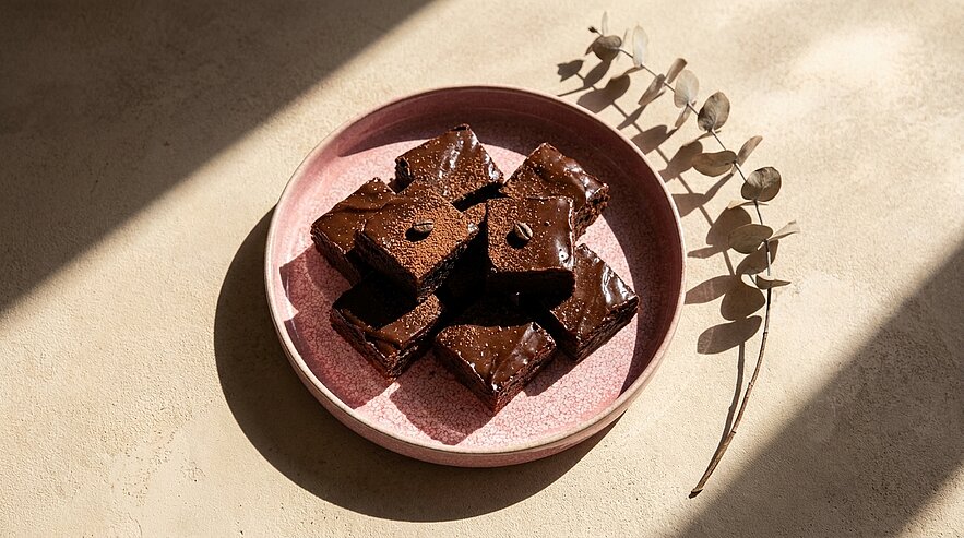 Mehrere quadratische, glänzend glasierte Schokoladenbrownies auf einem rosa Teller, angerichtet im warmen Sonnenlicht.