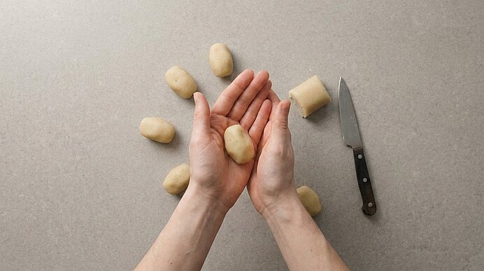 Zwei Hände rollen eine geschälte Kartoffel zu einer kleinen Kugel auf der Arbeitsfläche; mehrere geschälte Kartoffeln und ein Messer liegen daneben.