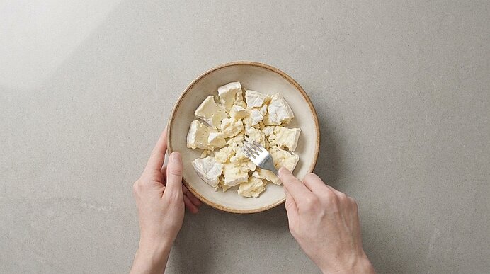 Zwei Hände halten eine Schale mit krümeligem weißen Käse; eine Gabel zerteilt die Käsestücke.