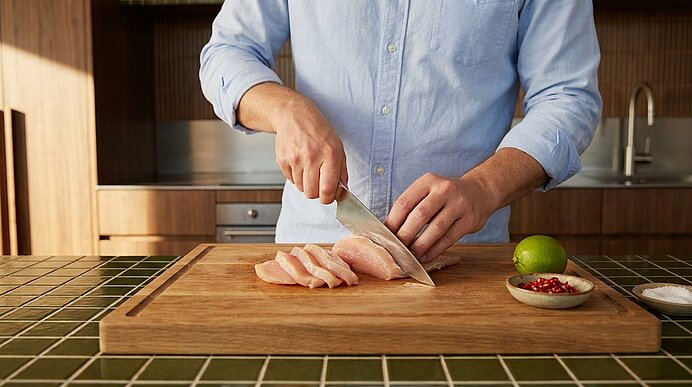 Hände einer Person schneiden rohes Hähnchenbrustfilet in dünne Scheiben auf einem Holzschneidebrett; daneben eine Limette und eine kleine Schale mit roten Gewürzen.
