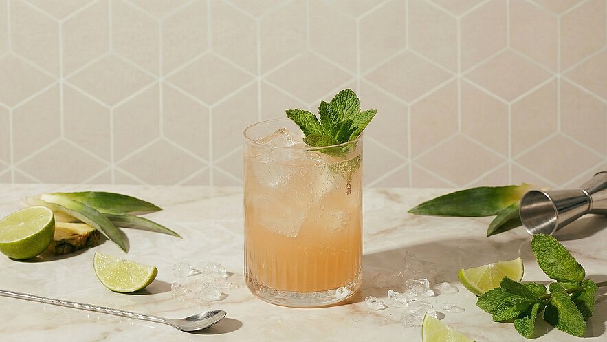 Ein Glas mit einem erfrischenden Getränk, gefüllt mit Eis, steht auf einer marmorierten Oberfläche. Frische Minze und Limettenscheiben dekorieren den Rand des Glases. Um das Glas liegen ein paar Limettenhälften und Eiswürfel, sowie ein Cocktailmessbecher aus Metall.