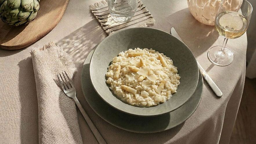 Ein Weißer Teller mit Risotto, belegt mit drei grünen Spargelspitzen und einem Blatt frischem Basilikum. Im Hintergrund sind kleine Schalen mit Butter und Parmesan sichtbar. Der Teller steht auf einem Holzbrett.
