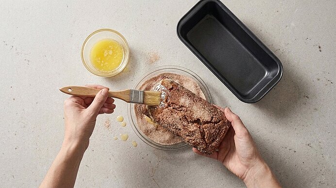 Zwei Hände halten ein frisch gebackenes Kastenbrot über einer Schüssel, während mit einem Pinsel geschmolzene Butter aufgetragen wird; eine leere Kastenform liegt daneben.