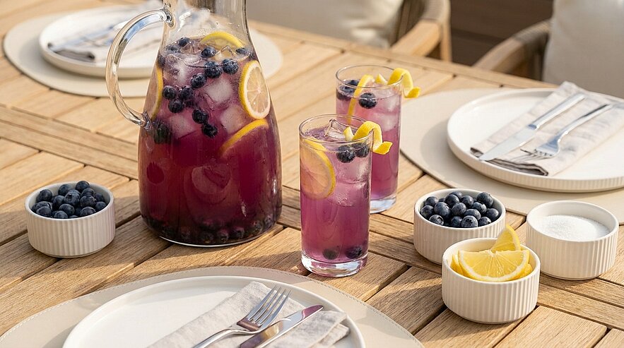 Auf einem Holztisch stehen eine Karaffe mit lila Getränk, zwei Gläser mit Eis und Zitronenscheiben sowie Schalen mit Heidelbeeren und Zitrusfrüchten. Die Szene vermittelt einladende Sommeratmosphäre.