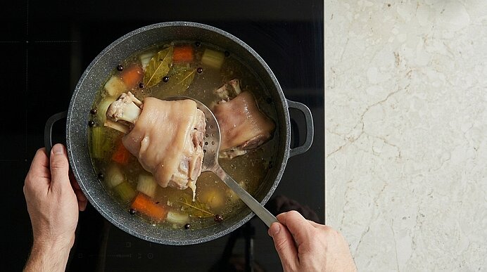 Person schöpft in einem Topf Brühe ein Stück Schweinehaxe; im Sud ziehen Gemüse (Karotten, Sellerie, Zwiebeln), Lorbeerblatt und Pfefferkörner.
