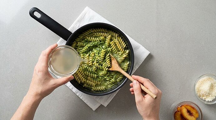 Zwei Hände gießen Nudelwasser zu Fusilli mit Pesto in einen Topf und rühren mit einem Holzlöffel um.