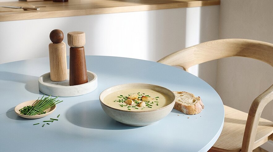 Schale mit cremiger Suppe, garniert mit Croutons und Schnittlauch; daneben ein Stück Brot, ein kleines Schälchen mit Schnittlauch und Holz-Salz‑ und Pfefferstreuer auf hellem Tisch.