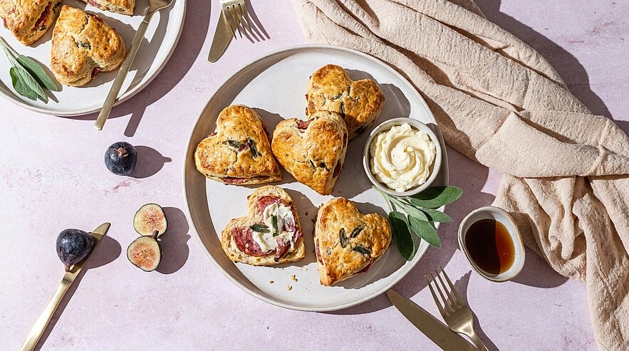 Auf einem Teller liegen herzförmige Scones, gefüllt mit Früchten und Frischkäse, garniert mit frischen Kräutern. Neben den Scones befindet sich eine Schale mit cremiger Sahne. Zwei halbierte Feigen und eine kleine Flasche Sirup sind ebenfalls zu sehen. Der Tisch ist in sanften Pastellfarben gedeckt.