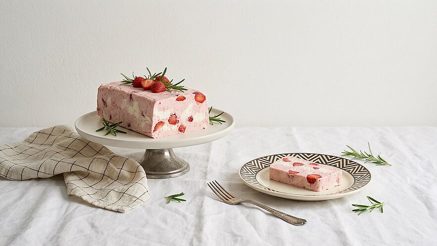 Ein eleganter Teller mit einem großen Stück Erdbeermousse, dekoriert mit frischen Erdbeeren und Rosmarin. Neben dem Hauptstück liegt ein auf einem Teller serviertes, kleineres Stück desselben Mousse. Der Hintergrund ist hell und schlicht, und die Oberfläche ist mit einem weißen Tuch bedeckt.