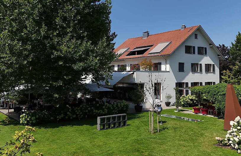 Ein stattliches, helles Gebäude mit einem roten Dach und weißen Wänden steht in einem gepflegten Garten. Im Freien sind Tische und Stühle auf einer Terrasse platziert, umgeben von grünen Pflanzen und Blumen. Sonnenlicht beleuchtet die Szene an einem klaren, blauen Himmel.