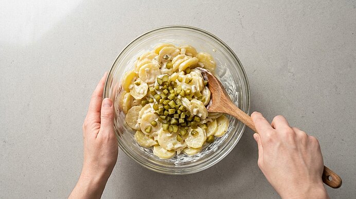 Zwei Hände rühren mit einem Holzlöffel Kartoffelsalat in einer Glasschüssel; Kartoffelscheiben mit cremigem Dressing und gewürfelten Gurken obenauf.
