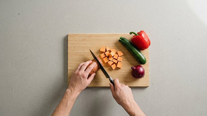 Hände schneiden auf einem Holzbrett eine Süßkartoffel in Würfel; daneben liegen eine rote Paprika, zwei Zucchini und eine rote Zwiebel.