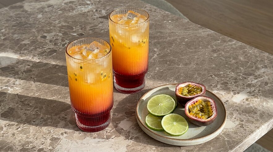 Zwei hohe, geschichtete Cocktails mit Eiswürfeln (orange-rot Verlauf) auf Marmoroberfläche; daneben ein kleiner Teller mit halbierten Passionsfrüchten und Limettenscheiben.