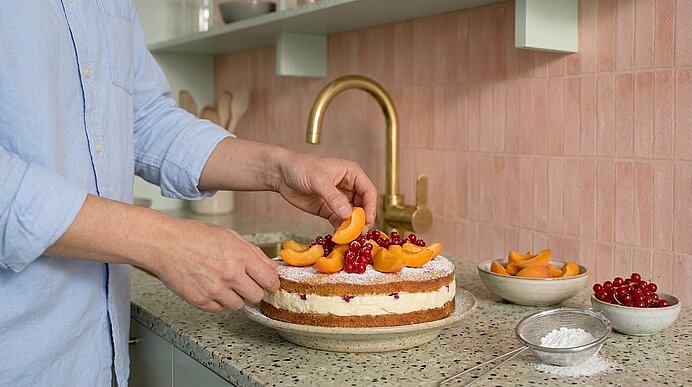 Hände dekorieren eine zweischichtige Torte mit Sahne, Puderzucker, Aprikosenhälften und roten Beeren auf einer Küchenarbeitsplatte.
