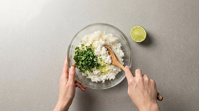 Zwei Hände rühren in einer Glasschüssel gekochten weißen Reis mit gehackten Kräutern (Koriander) und Limettensaft; halbierte Limette liegt daneben.