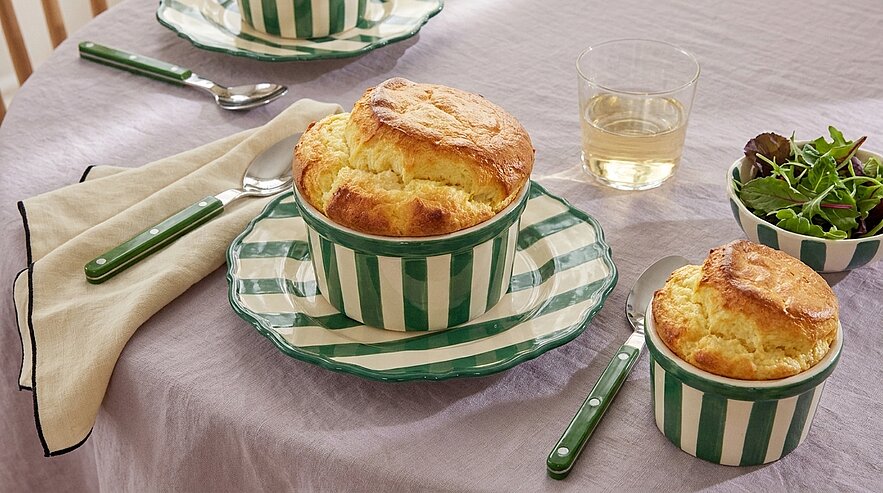 Zwei goldbraun aufgegangene Soufflés in grün‑weiß gestreiften Ramekins auf Tellern, dazu Besteck, ein Glas Wasser und eine kleine Schüssel Salat auf gedecktem Tisch.