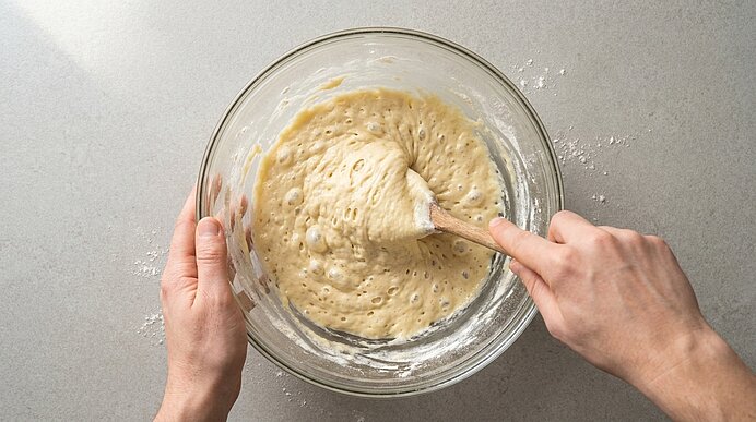 Zwei Hände rühren mit einem Holzlöffel einen blasenreichen, dickflüssigen Teig in einer großen Glasschüssel auf hellem Untergrund.