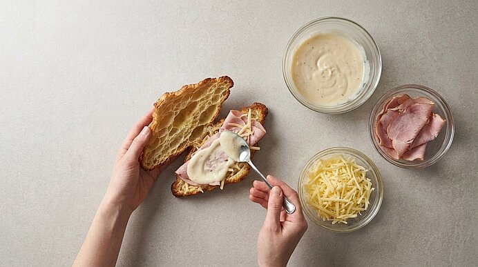 Zwei Hände verstreichen mit einem Löffel Béchamelsauce auf Schinken und Käse in einem aufgeschnittenen Croissant.