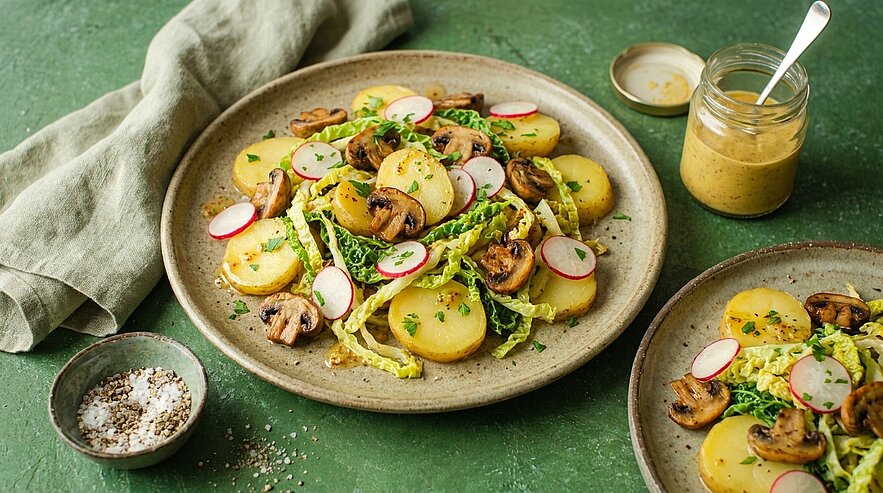 Teller mit Kartoffelsalat: dicke Kartoffelscheiben, gebratene Champignons, fein geschnittener Wirsing, Radieschenscheiben und Kräuter, daneben ein Glas Senfdressing.