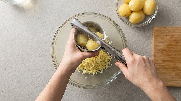 Zwei Hände pressen gekochte Kartoffeln mit einer Kartoffelpresse in eine Glasschüssel, in der sich geriebener Käse befindet.