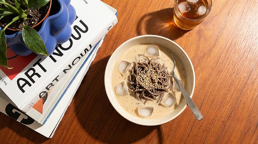 Eine Schüssel mit Soba-Nudeln in einer cremigen Sauce, garniert mit Eiswürfeln und Sesam. Daneben steht ein Glas mit einem kalten Getränk und im Hintergrund stapeln sich Kunstbücher. Ein kleiner Topf mit Pflanzen ergänzt die Szene.