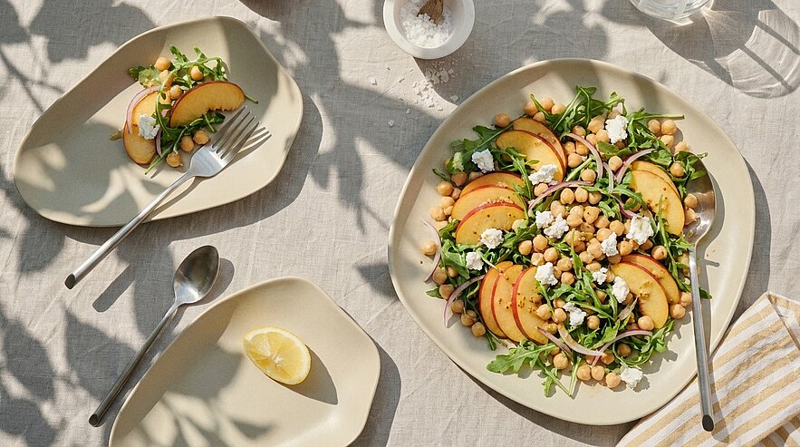 Große Servierplatte mit Rucola-Salat, Pfirsichscheiben, Kichererbsen und zerbröseltem Feta; daneben kleiner Teller mit Zitronenscheibe und Besteck.