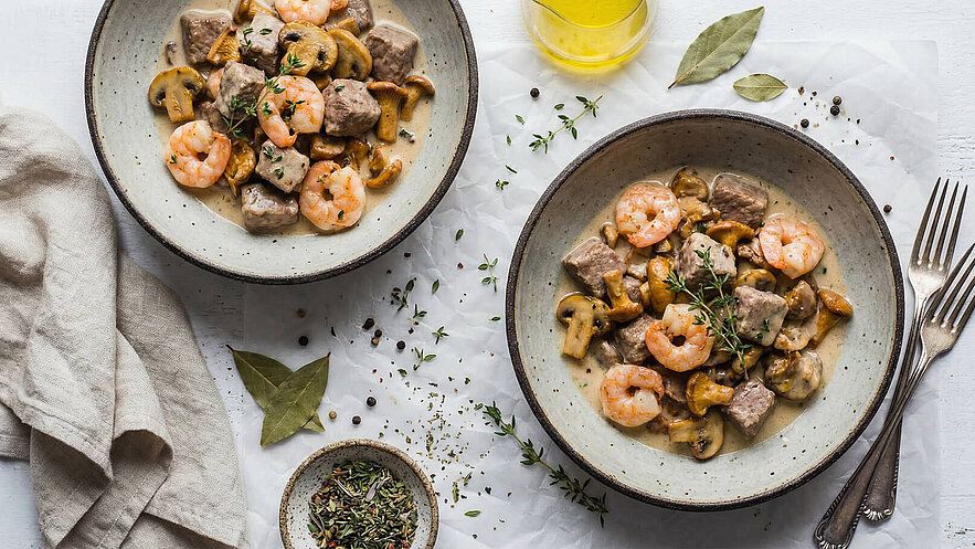 Zwei Schalen mit einem herzhaften Gericht aus Garnelen, Fleischstücken und Pilzen in einer cremigen Sauce. Frische Kräuter, wie Thymian, garnieren das Essen. Im Hintergrund sind ein Glas Öl und Gewürze auf einem Tisch sichtbar.