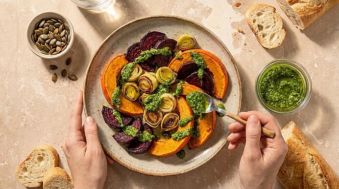 Hände halten einen Teller mit gerösteten Kürbisscheiben, Roter Bete und Lauchröllchen, beträufelt mit grüner Kräuterpesto.