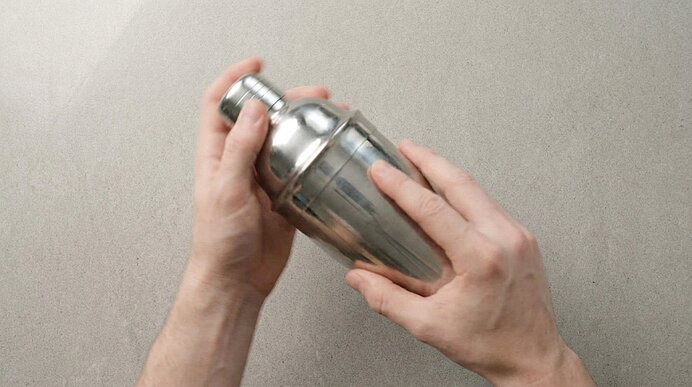 Zwei Hände schütteln einen geschlossenen Cocktailshaker aus Edelstahl über einer hellgrauen Arbeitsplatte.