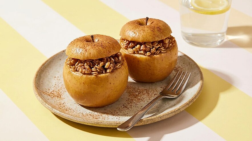 Zwei im Ofen gebackene, ausgehöhlte Äpfel, gefüllt mit knusprigem Nuss‑Granola, auf einem Teller mit Gabel.