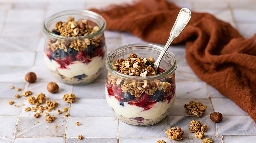 Zwei kleine Gläser mit geschichtetem Joghurt, Beerenkompott und knusprigem Granola; eines mit Löffel, verstreute Granola-Stücke und Haselnüsse.