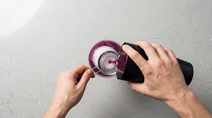 Zwei Hände gießen aus einem schwarzen Shaker einen schaumigen, violetten Smoothie in ein Glas; mit einem Löffel wird die Schaumkrone reguliert.