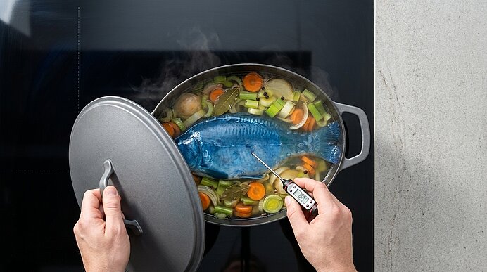 Draufsicht eines kochenden Topfes mit einem blauen ganzen Fisch auf einem Bett aus Gemüse; eine Hand hebt den Deckel, eine andere misst die Kerntemperatur mit einem Fleischthermometer.