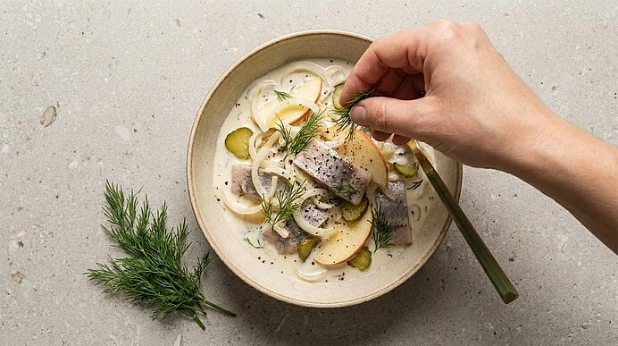 Schale mit cremigem Heringssalat: Heringstücken, Apfelscheiben, Gewürzgurken und Zwiebelringen in Sauce; Hand streut frischen Dill.
