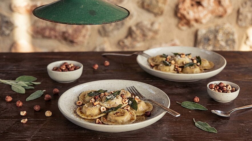 Zwei Teller gefüllte Ravioli mit brauner Butter, gerösteten Haselnüssen und Salbeiblättern auf rustikalem Holztisch; kleine Schälchen mit Haselnüssen daneben.