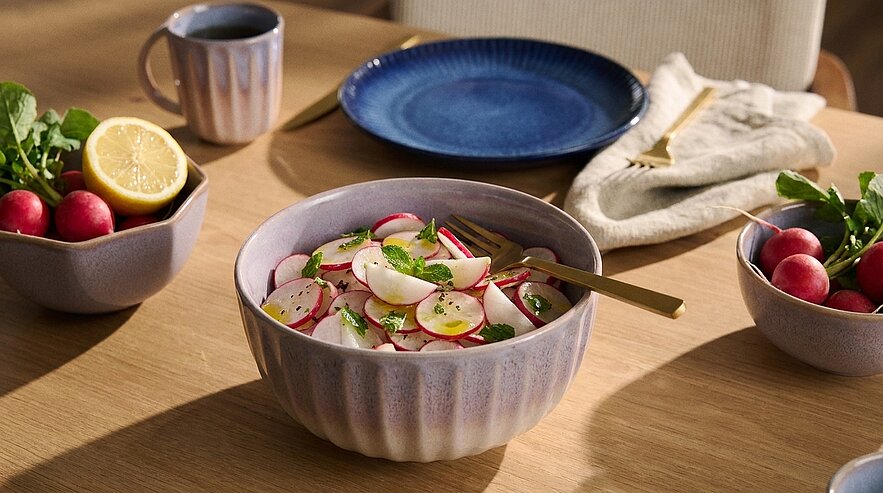 Keramikschüssel mit frischem Radieschensalat: dünn geschnittene Radieschen, Kräuter und Dressing auf einem Holztisch, daneben Schälchen mit ganzen Radieschen.