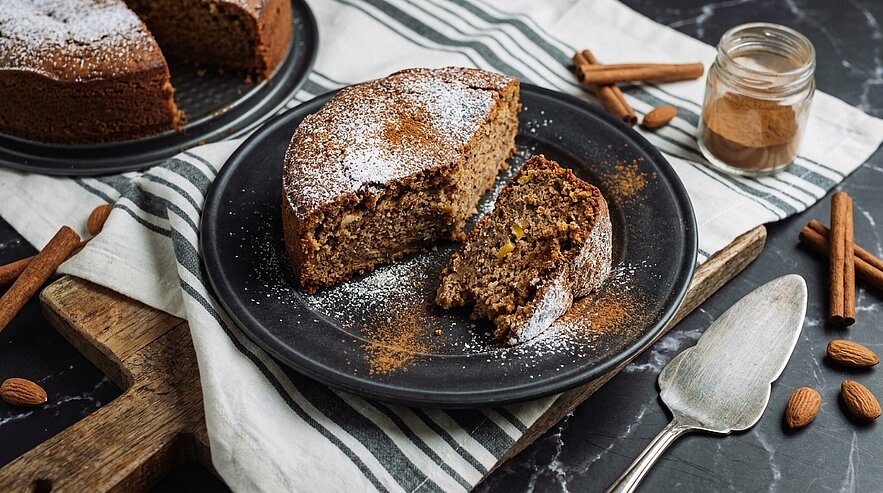 Ein saftiger, gewürzter Kuchen auf einem dunklen Teller, mit einer bereits angeschnittenen Portion. Der Kuchen ist mit Puderzucker bestäubt und umgeben von Zimtstangen und Mandeln. Im Hintergrund steht ein Glas mit Zimt. Der Tisch hat eine gestreifte Tischdecke.