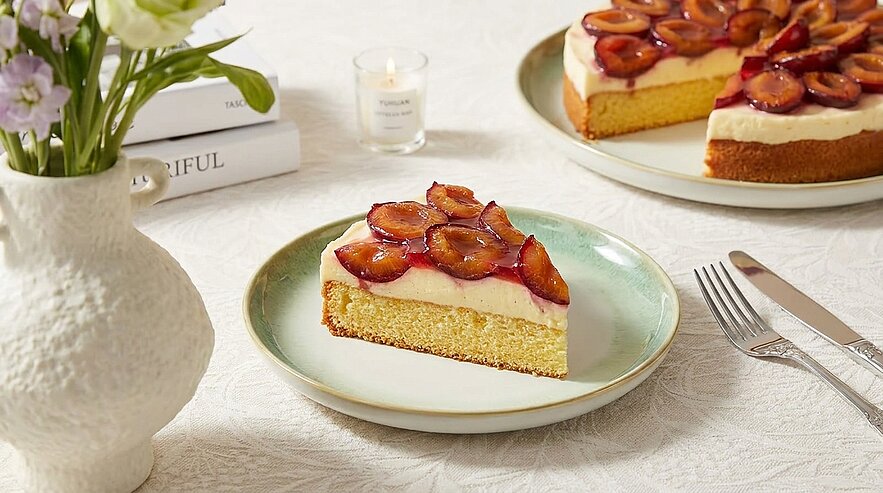Ein Stück Kuchen mit einer Schicht Sahne und karamellisierten Früchten, auf einem weißen Teller serviert. Daneben steht eine Torte mit ähnlicher Ausstattung. Im Hintergrund steht eine Vase mit frischen Blumen.