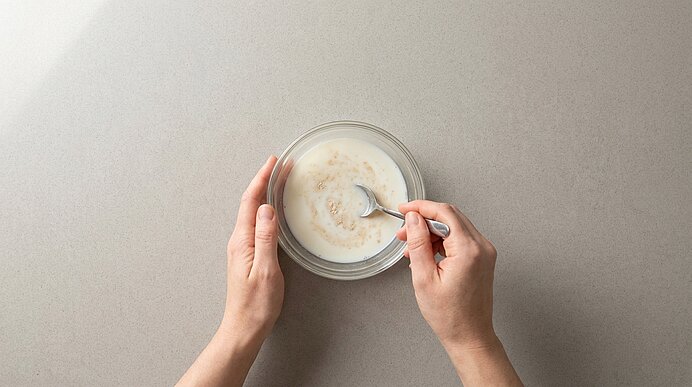 Vogelperspektive: Hände halten eine Glasschüssel, in der mit einem Löffel Haferflocken in Milch verrührt werden.