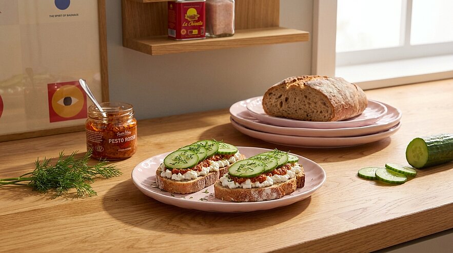 Zwei offene Vollkornbrote auf einem Teller mit Frischkäse, Tomatenstückchen, Dill und Gurkenscheiben; daneben ein Laib Brot, Gurke und ein Glas Aufstrich auf einer Holzarbeitsplatte.