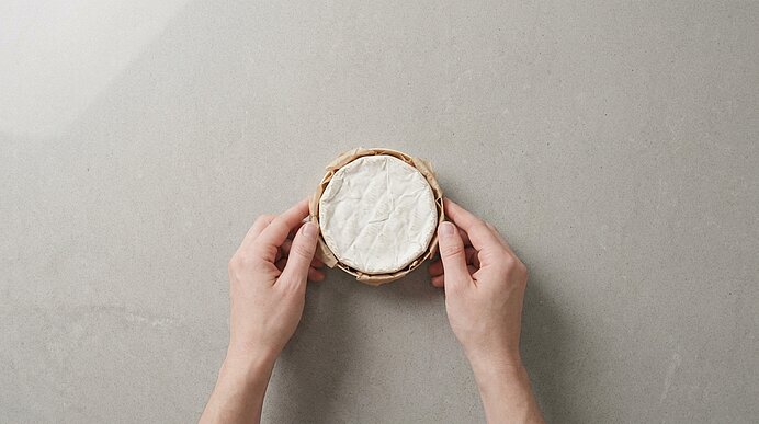 Zwei Hände halten einen runden Weichkäse (Camembert/Brie) in einem kleinen Korb auf grauem Hintergrund.