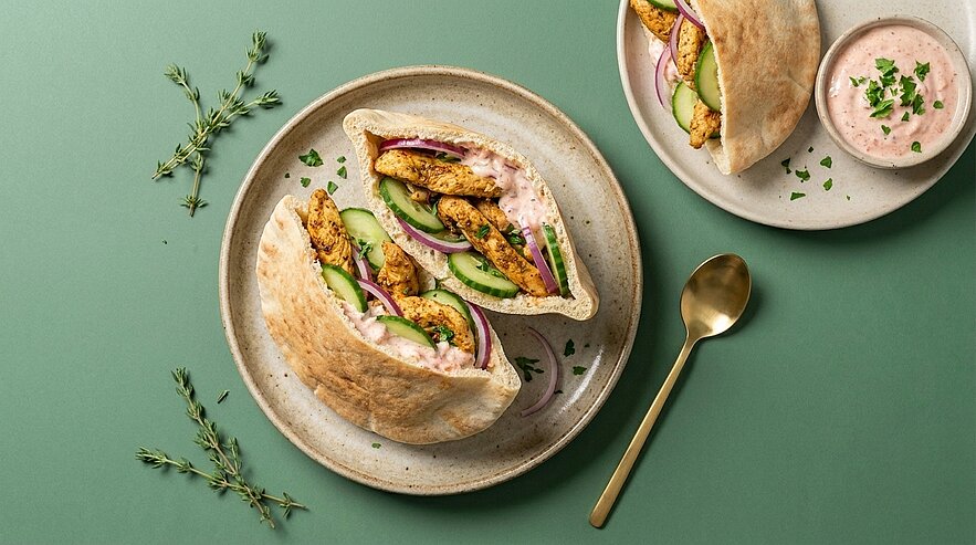 Zwei Teller mit gefüllten Pita-Taschen, gefüllt mit gegrillten Hähnchenstreifen, Gurkenscheiben, roten Zwiebelringen und cremiger rosa Kräutersauce; daneben kleine Schale mit Sauce und goldener Löffel.