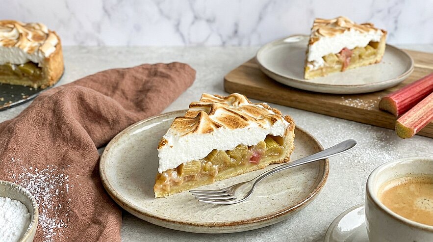 Ein Stück Rhabarberkuchen mit einer luftigen Baisertorte auf einem Teller, daneben ein Teelöffel. Im Hintergrund befinden sich weitere Stücke Kuchen sowie eine Tasse Kaffee. Die Oberfläche des Kuchens ist goldbraun und leicht karamellisiert.