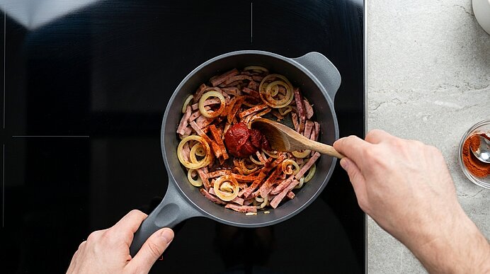 Zwei Hände rühren mit einem Holzlöffel Zwiebelringe, Schinkenstreifen und Tomatenmark in einer Pfanne auf dem Herd.