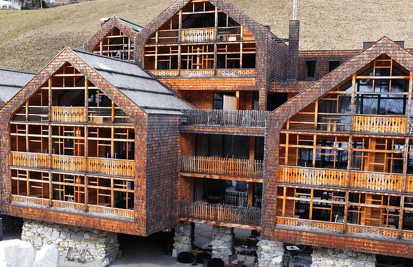 Mehrteilige hölzerne Bergchalet-Anlage mit mehreren Giebeldächern, durchgehenden Holzbalkonen, großen Fenstern und steinernem Sockel vor einem grasigen Hang.